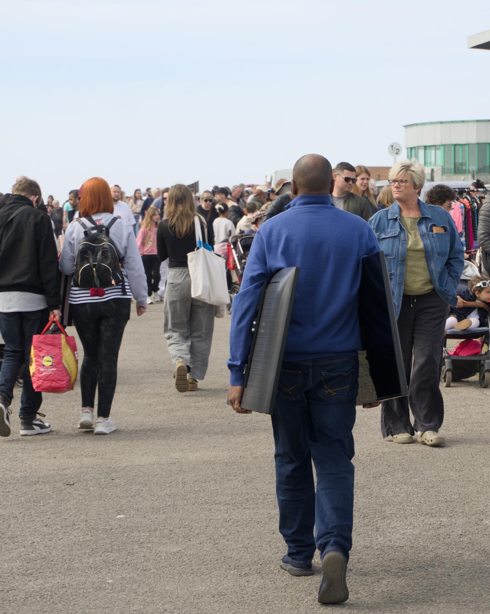 A man in a blue jumper walking away through a busy seafront market carrying a large CRT television under his arm, with crowds of shoppers and stalls in the background