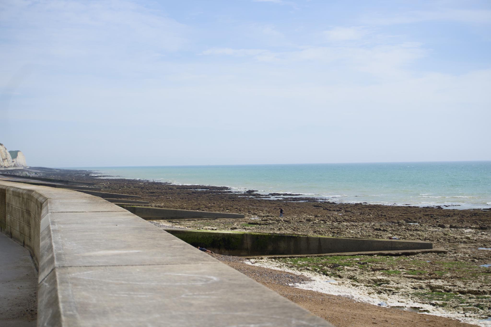 A long curved concrete sea wall stretching towards distant white chalk cliffs, with a rocky low-tide beach and the sea on the right under a hazy blue sky