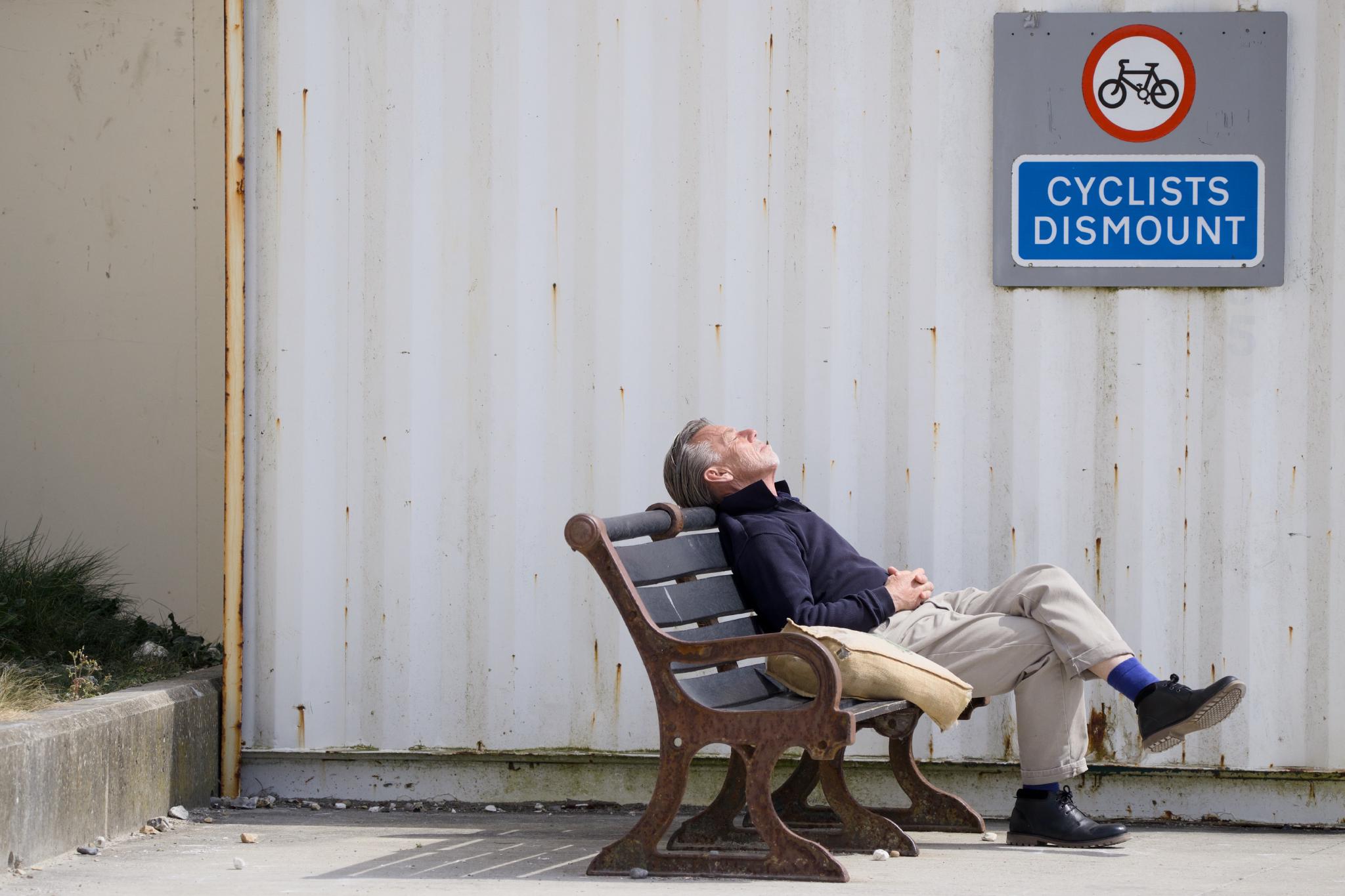 An older man in a navy jumper, beige trousers and blue socks reclining on a wooden bench, head tilted back to the sun, beneath a 'CYCLISTS DISMOUNT' sign on a corrugated white wall