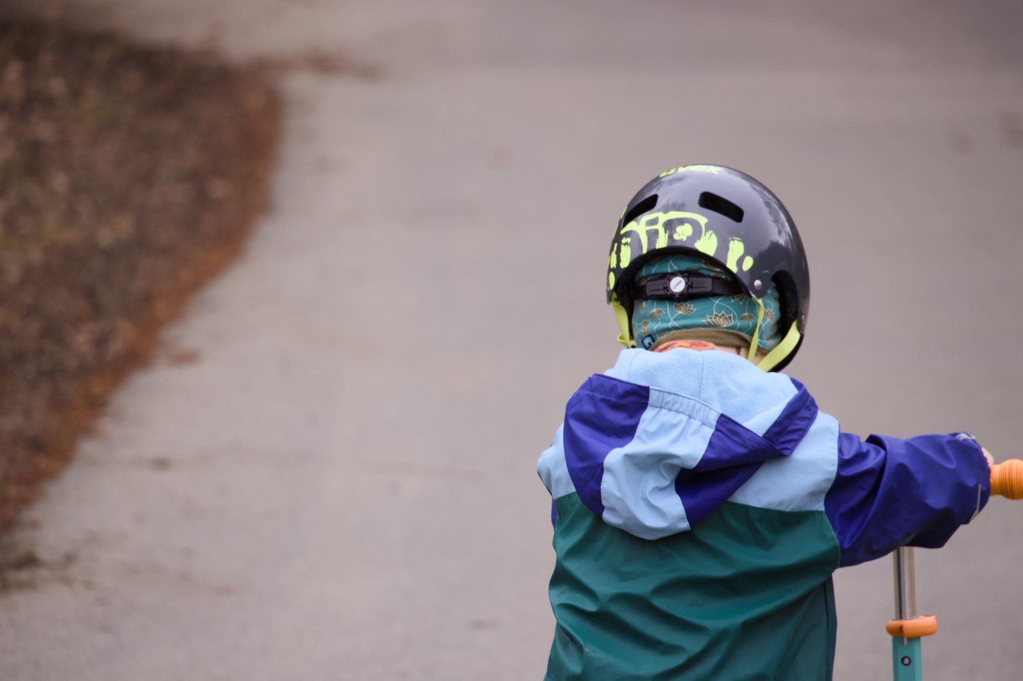Small child from behind on a balance bike, wearing a dark helmet and blue rain jacket